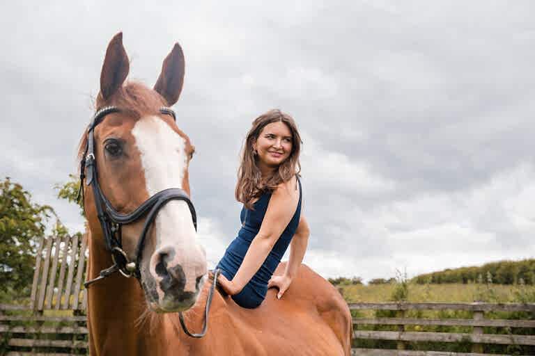 A woman in a blue dress sits on a brown horse outdoors with a fence and cloudy sky in the background.