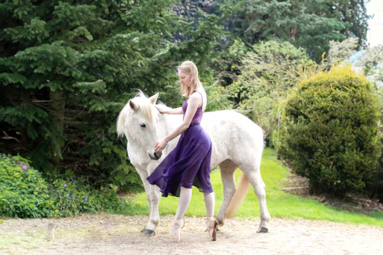 A woman wearing a purple dress and ballet shoes performs a dance move with a white horse standing beside her in a garden setting.