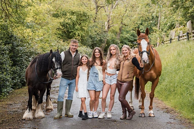 A group of five people, consisting of three girls and two adults, stands on a tree-lined path with two horses.