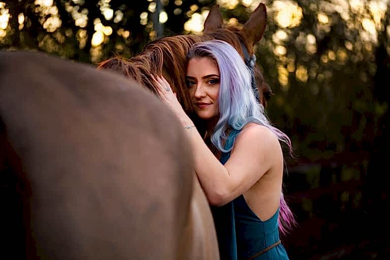 A woman with pastel-colored hair embraces a brown horse while standing outdoors, surrounded by greenery and a wooden fence.