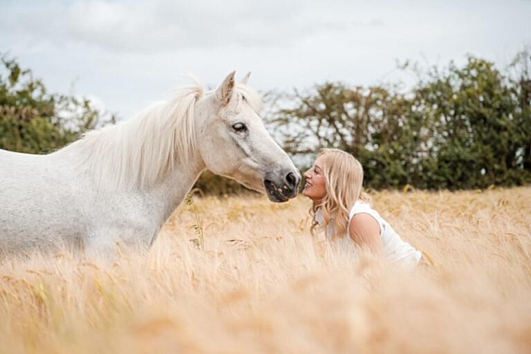 A blonde woman kneels in a field of tall grass, affectionately touching noses with a white horse.