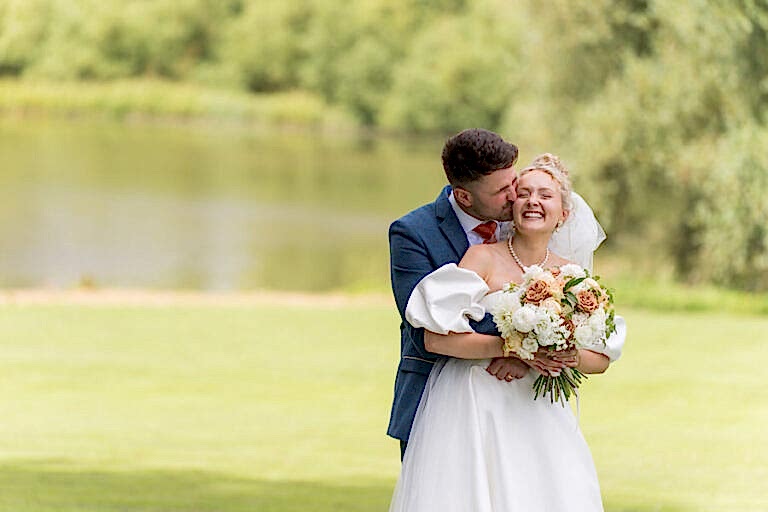 A bride and groom embrace in front of a lake, with the groom kissing the bride on the cheek, both smiling. The bride holds a bouquet of flowers and the background features greenery and water.