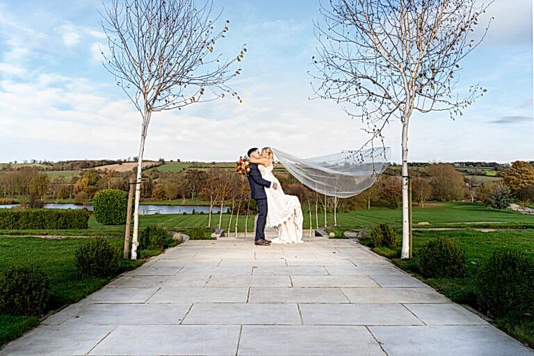Bride and groom share a kiss outdoors, with the groom lifting the bride. Her veil flows behind as they stand on a stone path, surrounded by trees and a scenic landscape.